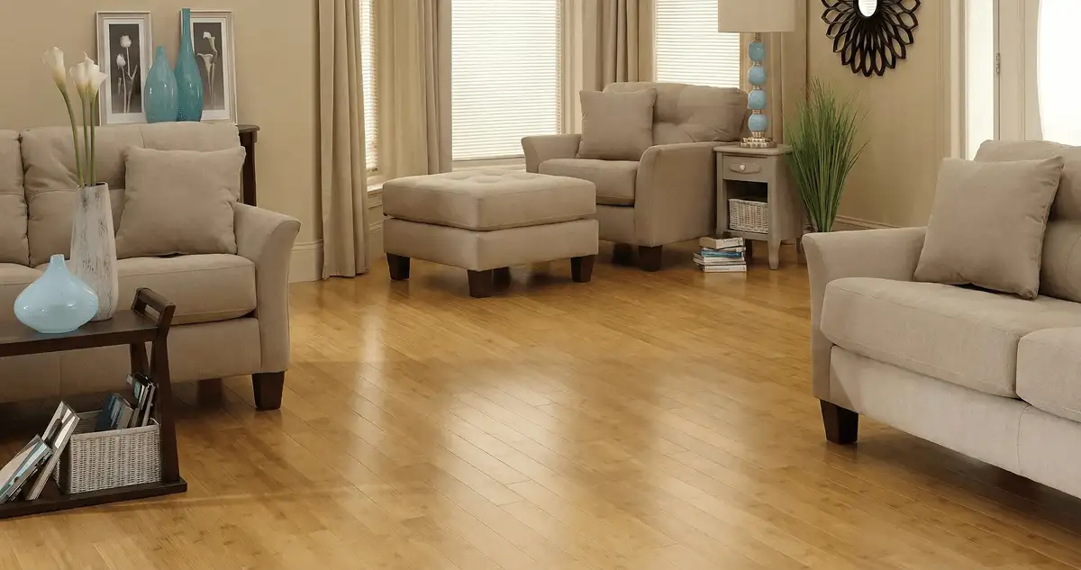 A living room with professionally installed bamboo flooring, beige sofas, a matching ottoman, a side table with a blue lamp, vases, framed pictures, and large windows with blinds and curtains in Jackson, MS